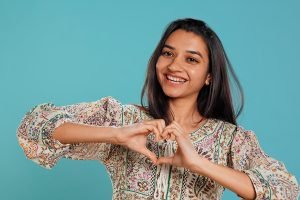 Portrait of smiling loving woman doing heart symbol shape gesture with hands, showing kindness. Happy affectionate girlfriend doing tender love gesturing for boyfriend, studio background, camera B